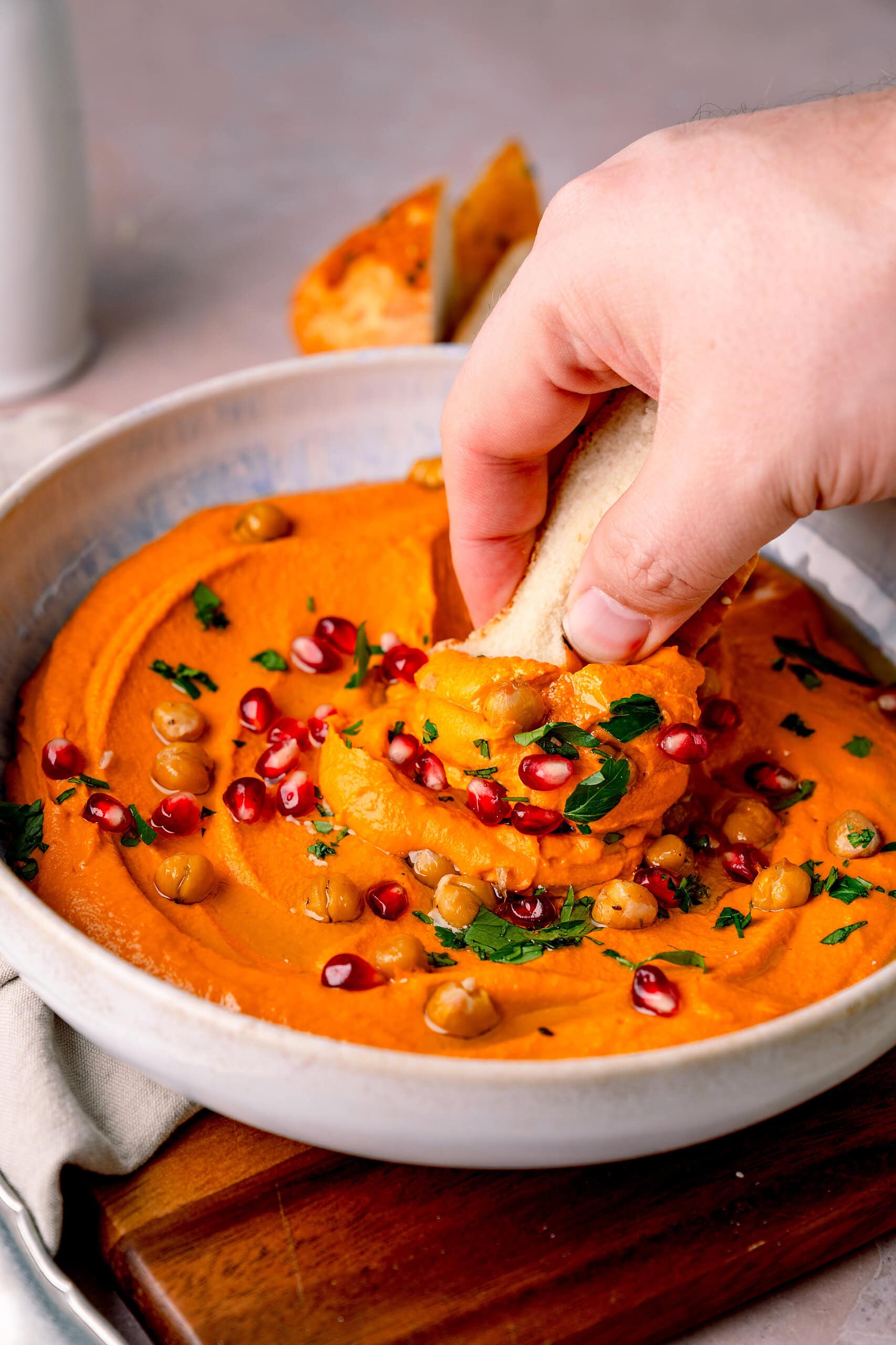 Roter Paprikahummus mit Kichererbsen, Granatapfel und Petersilie, eine Hand, ein Stück Brot, das sich etwas vom Dipp nimmt